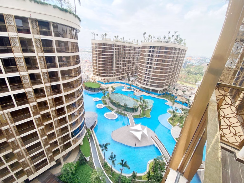 View from a balcony at Orkide The Royal, overlooking modern, curved high-rise buildings surrounding a large, irregularly shaped swimming pool with islands, walkways, palm trees, and landscaped green areas.