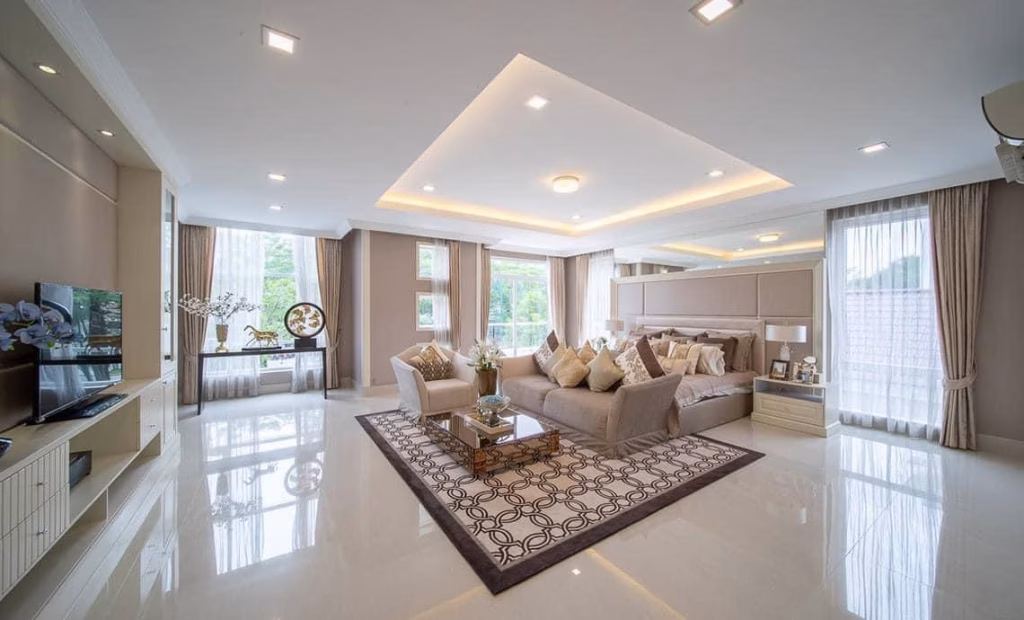 Spacious, modern bedroom in a house for sale, featuring large windows, neutral-toned decor, a king-sized bed with many pillows, patterned rug, coffee table, and flat-screen TV; natural light fills the elegant room.