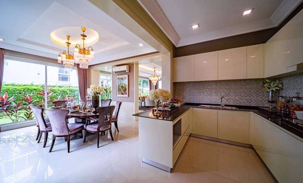 Modern kitchen and dining area in this house for sale features cream cabinets, black countertops, and elegant chandeliers. Large windows fill the space with natural light, showcasing a landscaped garden. The dining table is beautifully set with flowers.