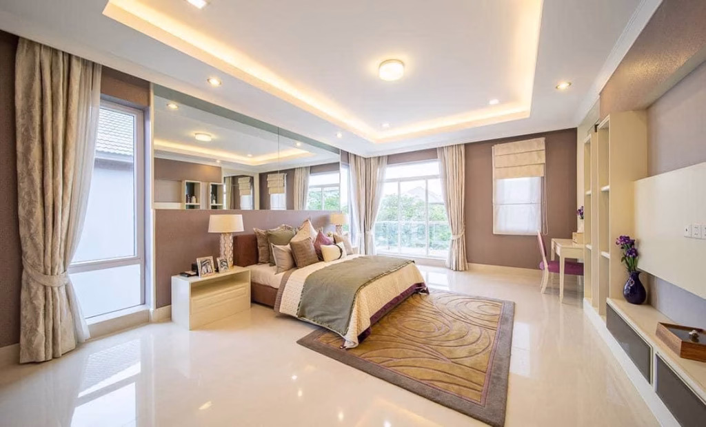 Spacious, modern bedroom in this house for sale features a large bed, neutral tones, mirrored wall, desk, built-in shelves, large windows with curtains, and recessed ceiling lighting for a bright and elegant atmosphere.