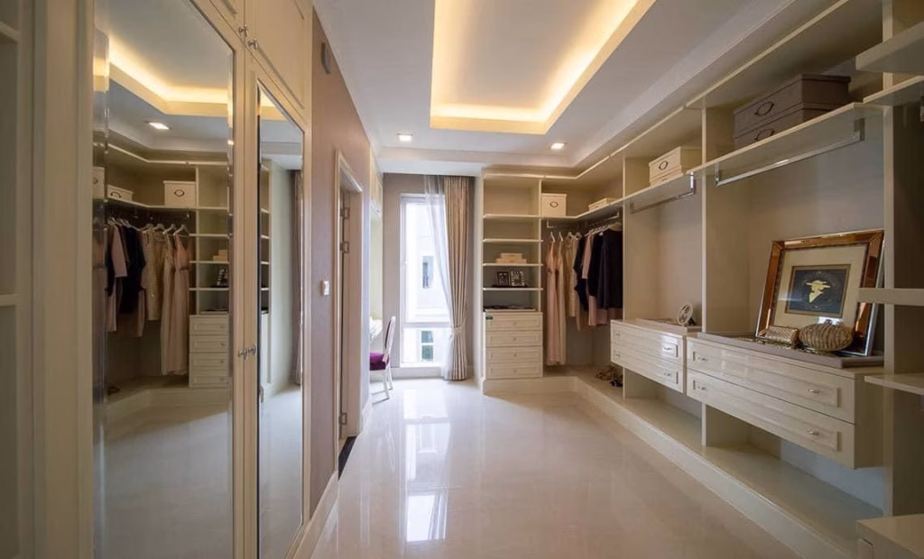 Spacious, modern walk-in closet in this house for sale, featuring hanging clothes, drawers, shelves, a large mirror, soft ceiling lights, decorative items, and a window at the far end letting in natural light.