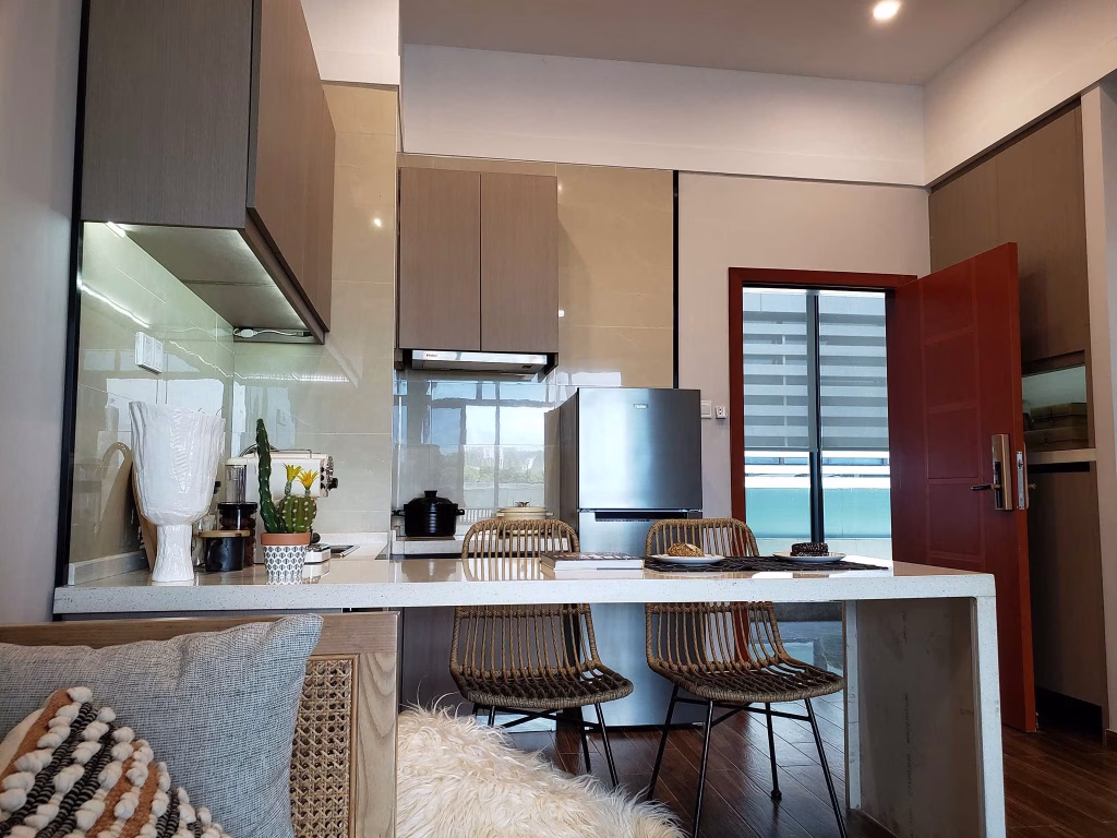 Modern kitchen in a Sihanoukville Studio For Sale features gray cabinets, a stainless steel refrigerator, and a breakfast bar with three wicker chairs. Countertop displays decor items and a coffee maker. A wooden door opens to natural light.