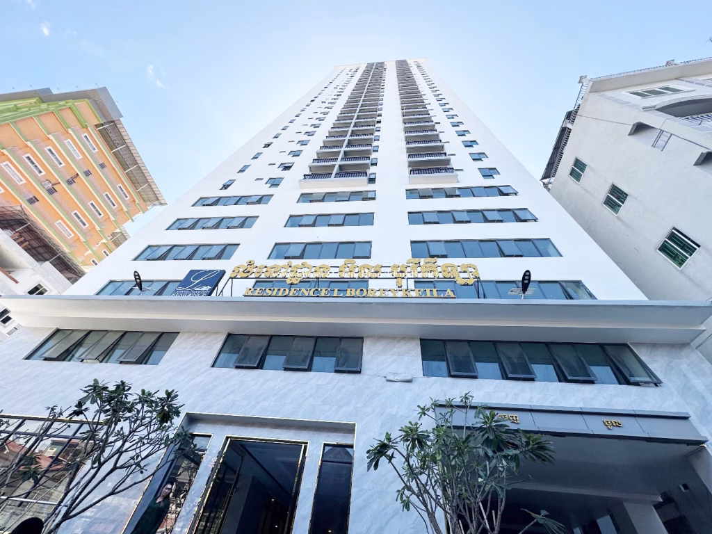 Tall white high-rise building seen from ground level, with a blue and gold RESIDENCE L BOSETTA VILLA sign above the entrance. This 1 bed condo is flanked by trees and neighboring buildings under a clear sky.
