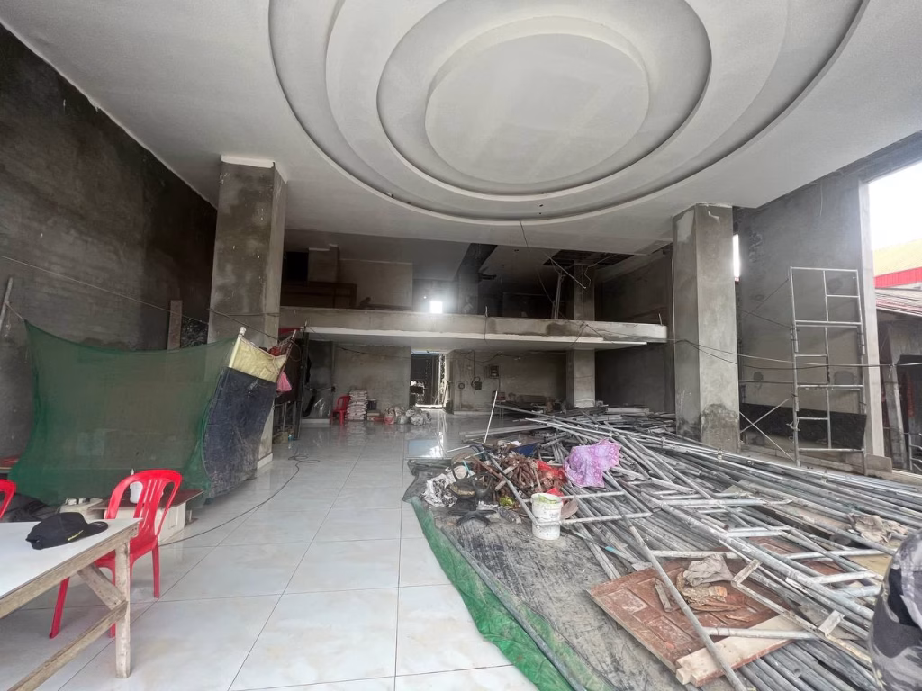 A large room under construction in a prime office building, with metal rods, tools, and scaffolding scattered on the tiled floor. Red plastic chairs and tables are on the left, and the ceiling features a circular recessed design.