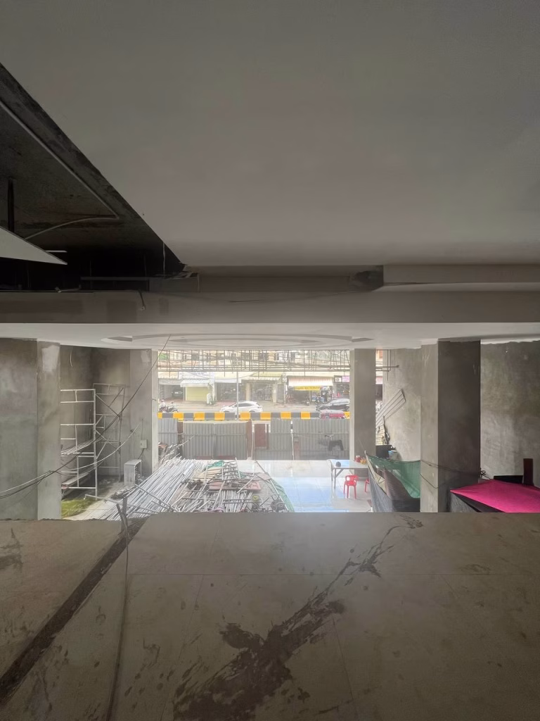 A view from inside a prime office building under construction, with scaffolding, poles, and materials visible. Large windows reveal traffic, barriers, and nearby buildings. Some chairs and tables are scattered around.