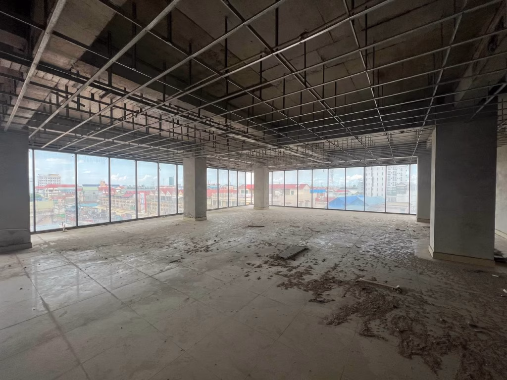 Large, empty room in a prime office building with an unfinished ceiling grid, dirty floor, and floor-to-ceiling windows overlooking a cityscape and cloudy sky. Natural light fills the spacious area.