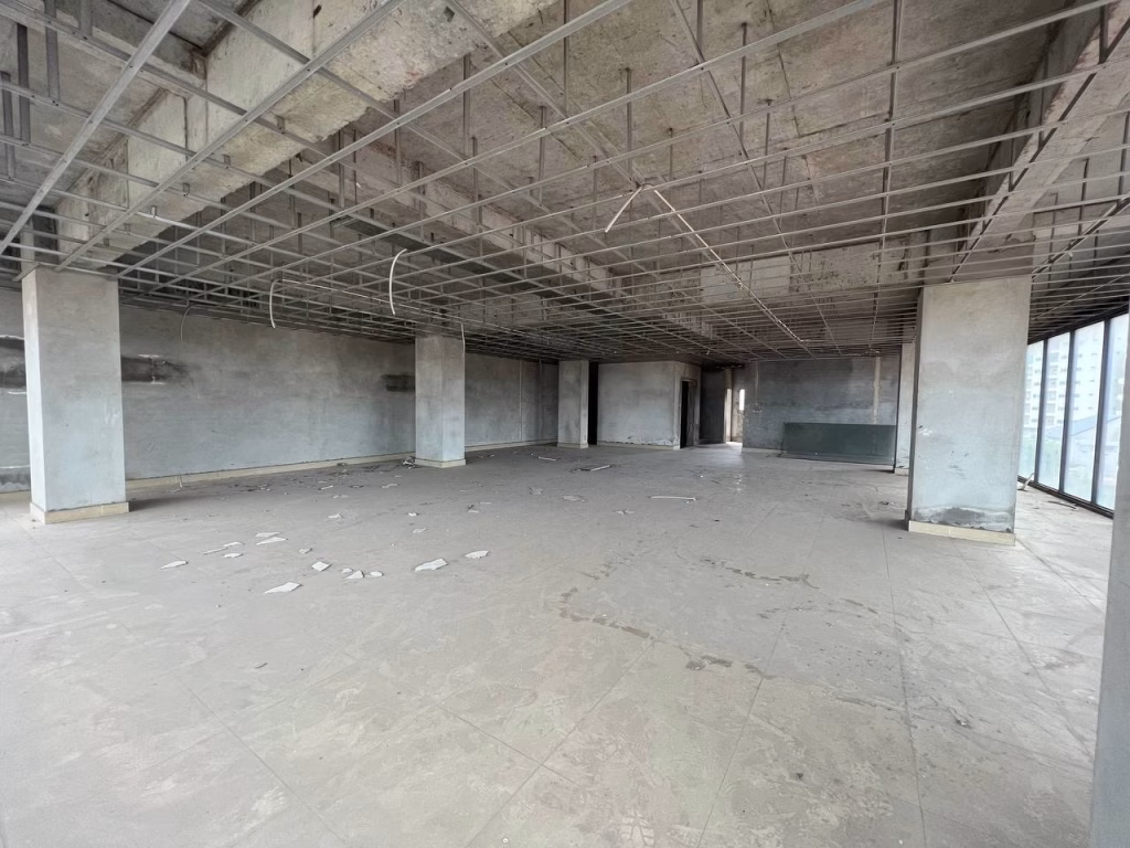 An empty, unfinished room in a prime office building with concrete walls, exposed ceiling grids, columns, and large windows on the right side. The dusty floor is scattered with debris and construction materials.