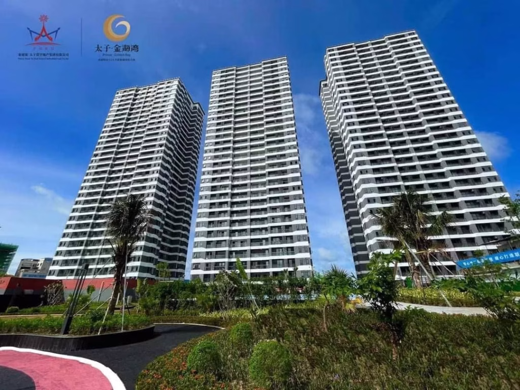 Three tall modern apartment buildings with white balconies stand side by side, surrounded by landscaped greenery and palm trees under a clear blue sky—perfect for anyone seeking a Sihanoukville Studio For Sale. Logos and Chinese text appear in the top left corner.
