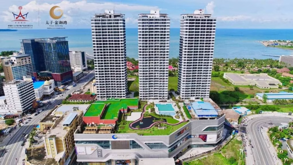 Aerial view of a modern coastal cityscape featuring three tall white residential towers, a shopping complex with rooftop gardens and pools, and curving roads near the ocean—perfect for those seeking a Sihanoukville Studio For Sale.