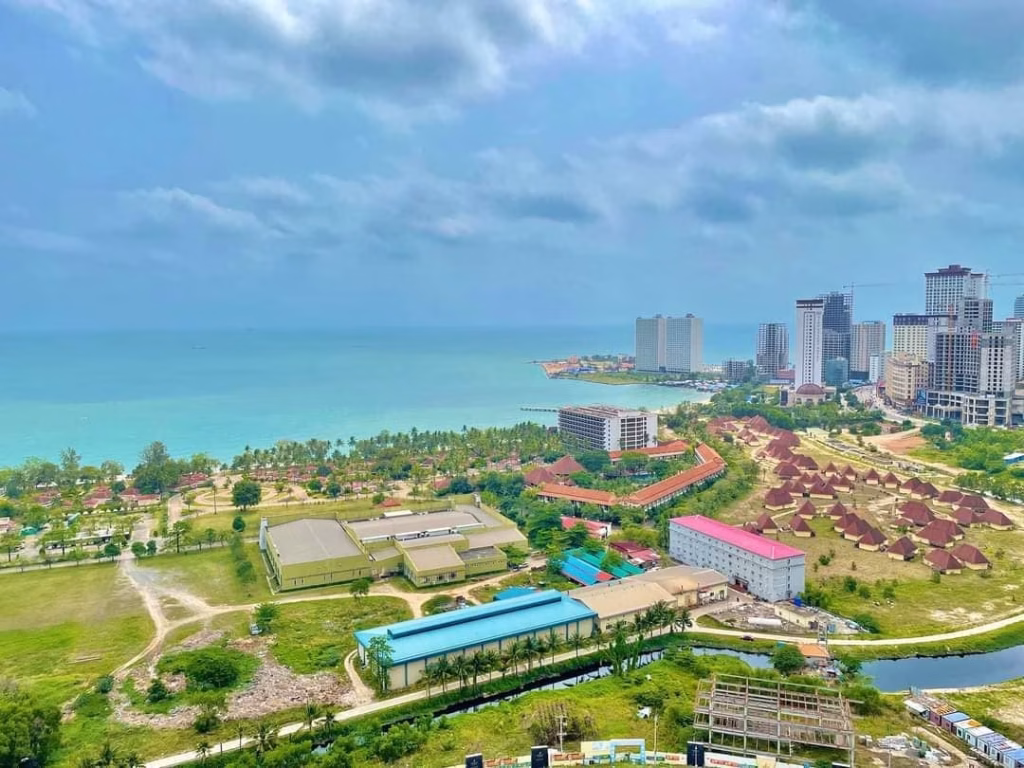 A coastal cityscape with tall buildings near the shoreline, green areas, scattered small structures, and clear blue ocean under a partly cloudy sky—an ideal spot to find your Sihanoukville studio for sale.