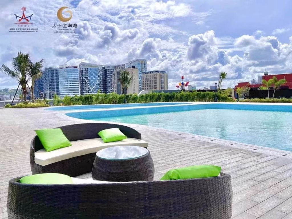 Modern outdoor pool area with round wicker couches and green cushions set on a paved deck. High-rise buildings, palm trees, and a partly cloudy sky form the backdrop—perfect for relaxing at your Sihanoukville Studio For Sale.
