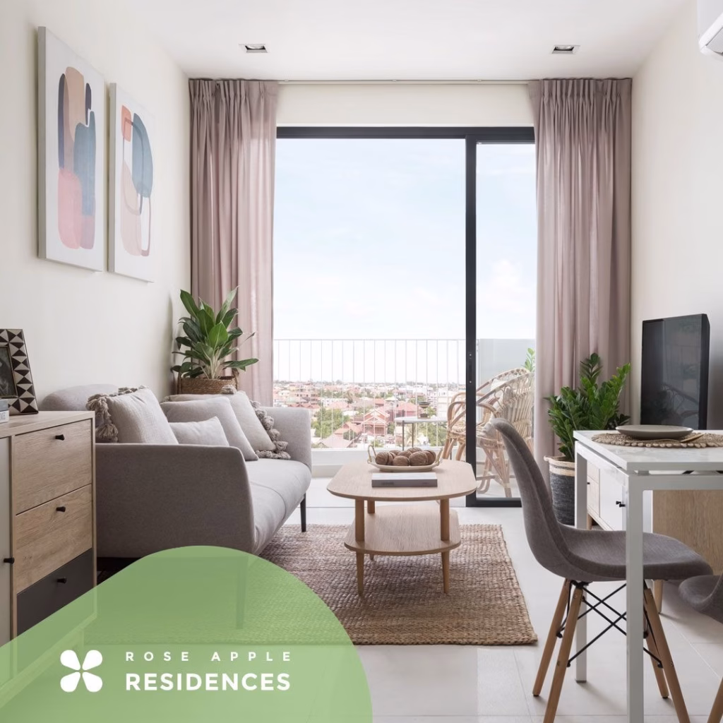 A modern, bright living room at Rose Apple Square features a gray sofa, coffee table, and a desk facing a computer monitor. Large glass doors open to a balcony with city views. Green plants and abstract art decorate the space. “Rose Apple Residences” logo is visible.