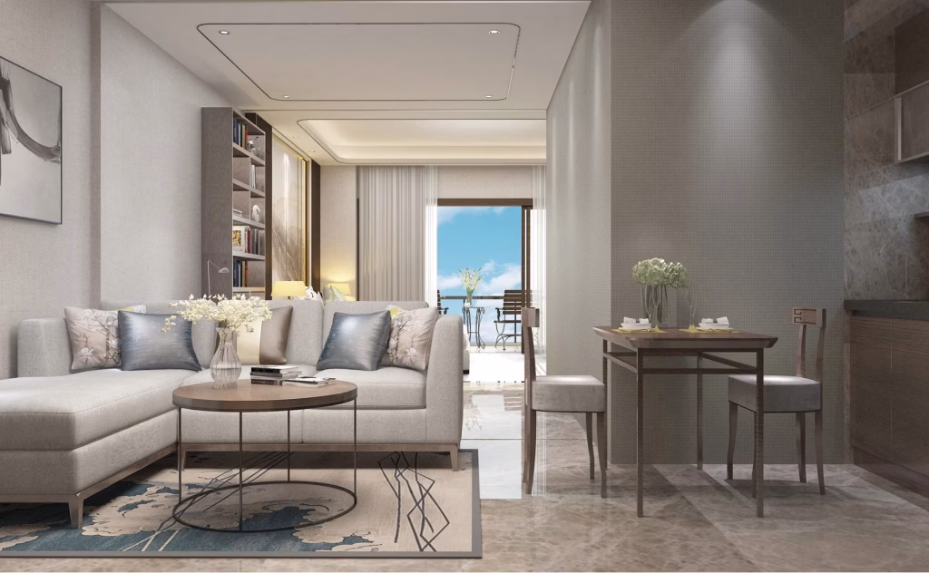 Modern living room at The Seagate Apartment features a light gray sectional sofa, round coffee table, and floral decor beside a cozy dining area for two. Large windows open to a balcony with sea views and blue sky.