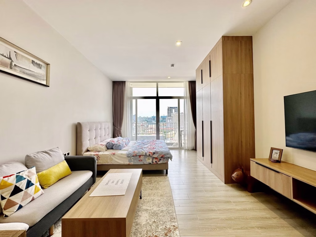 A modern serviced apartment 1 bedroom features a bed, sofa, wooden coffee table, TV stand, and large wardrobe. Floor-to-ceiling windows offer a city view, while neutral decor and light wood furniture create a bright, airy feel.