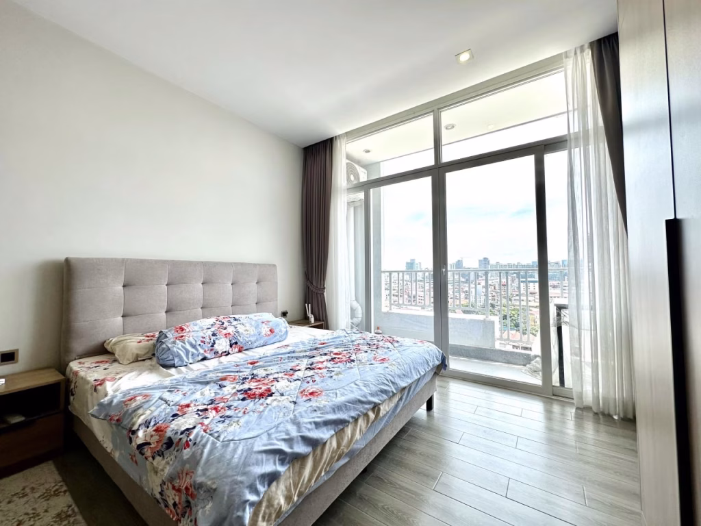 A bright, modern serviced apartment 1 bedroom with a large bed covered in floral bedding, light wood flooring, and floor-to-ceiling windows leading to a balcony with a city view. White curtains frame the windows, letting in natural light.