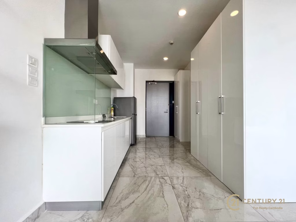 Modern kitchen in a studio apartment for rent, featuring glossy white cabinets, marble floor, built-in stove, and a stainless steel refrigerator. A black door is at the end of the hallway, with CENTURY 21 logo in the bottom right corner.