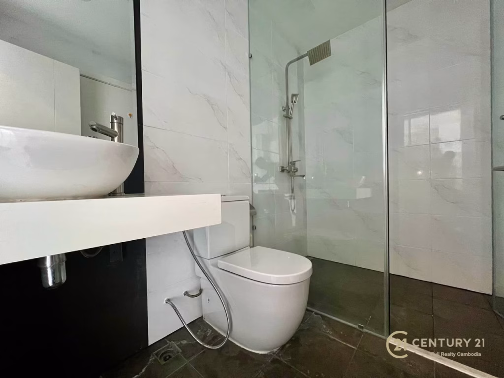 Modern bathroom in a studio apartment for rent, featuring a white sink on a floating counter, toilet, and glass-enclosed shower with rain showerhead. White tiled walls and dark floor. Century 21 Realty Cambodia logo is visible.