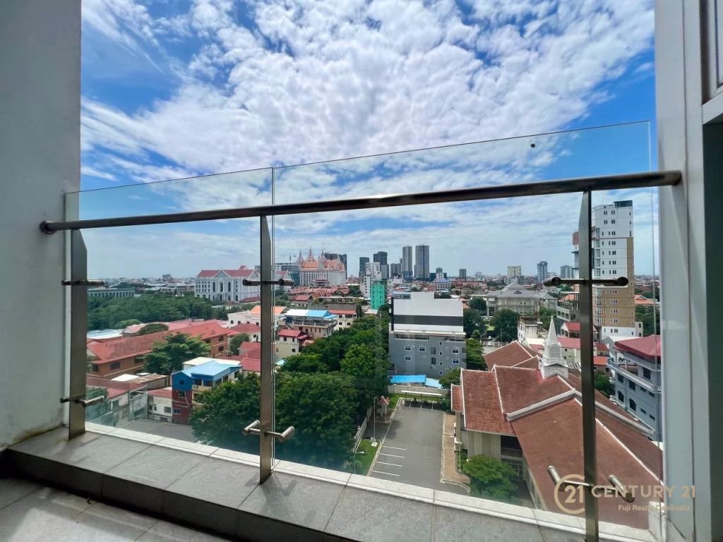 View from a balcony with a glass railing overlooking a cityscape of mixed low-rise and high-rise buildings under a partly cloudy sky—perfect for those seeking a studio apartment for rent. The CENTURY 21 logo is visible in the bottom right corner.