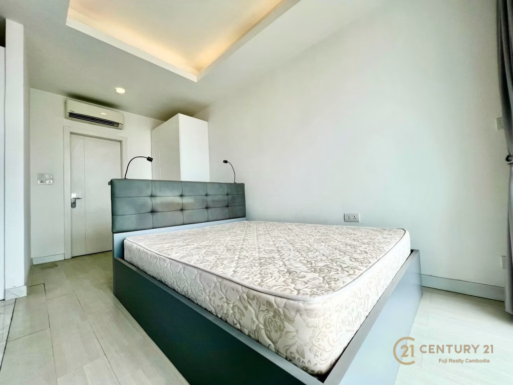 Minimalist bedroom in a studio apartment for rent, featuring a large bed and mattress, white walls, built-in headboard with reading lights, air conditioning unit, and modern recessed ceiling lighting. Century 21 logo in the corner.