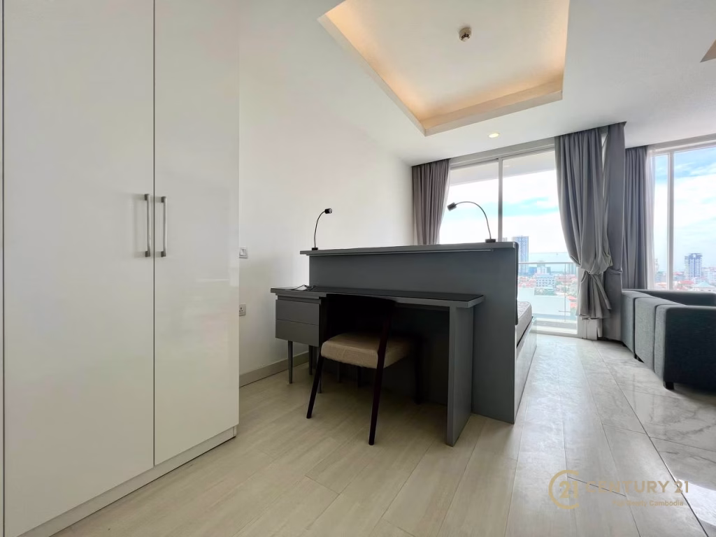 Modern, bright studio apartment for rent featuring a bed near a large window, a small desk and chair at the foot, white wardrobe on the left, light wood flooring, and city views through floor-to-ceiling windows.
