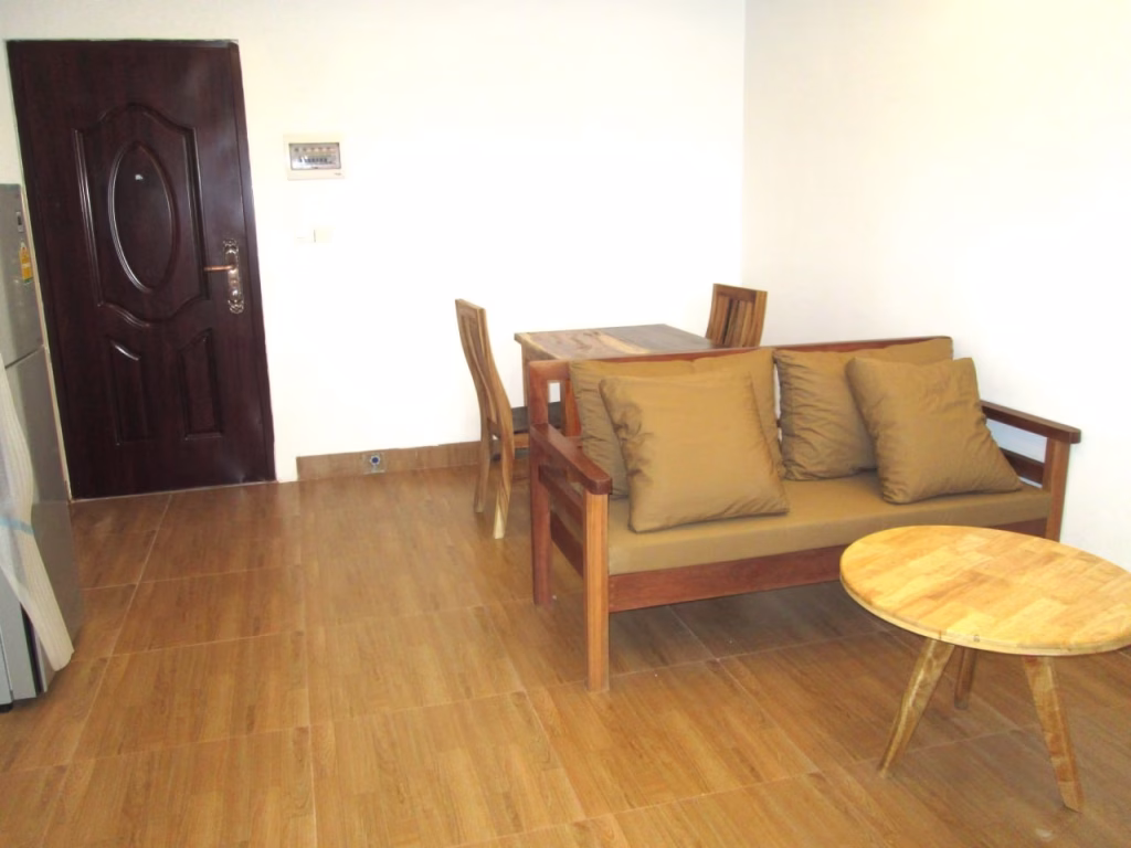 A cozy studio apartment for sale featuring a small living and dining area with a wooden sofa with tan cushions, matching dining table and chairs, a round coffee table, light brown tiled floor, and a dark brown door.