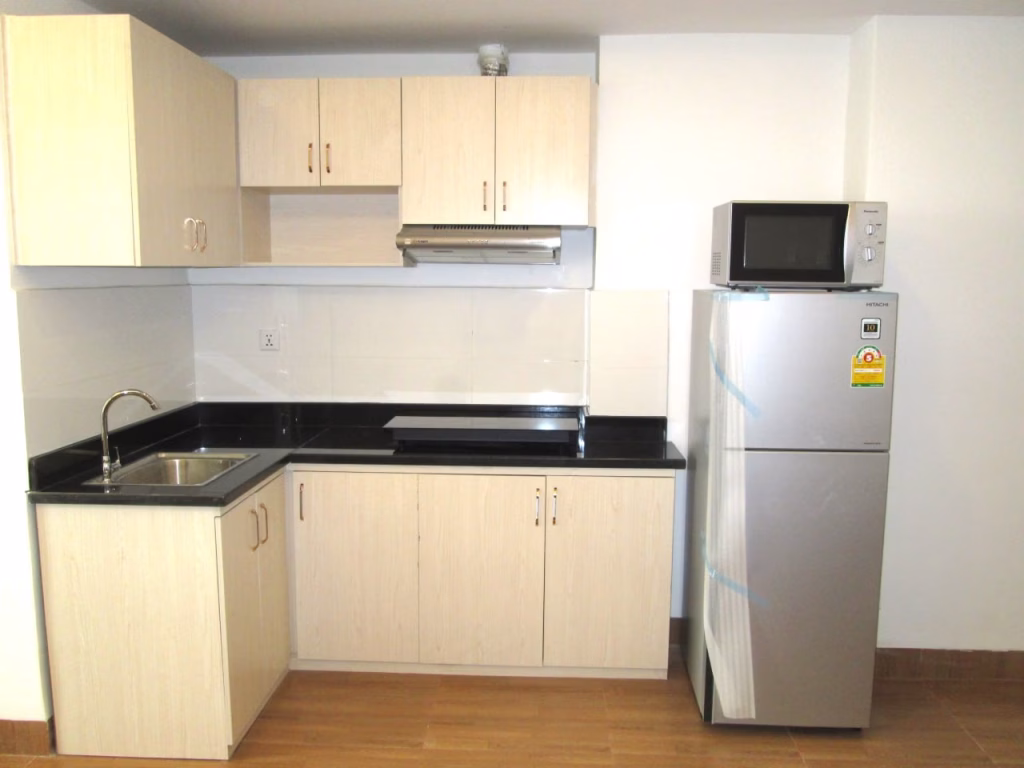 A compact kitchen in a studio apartment for sale features light wood cabinets, a black countertop, a stainless steel sink, a flat cooktop, and a silver refrigerator with microwave. The wooden floor and white walls create a bright, modern feel.