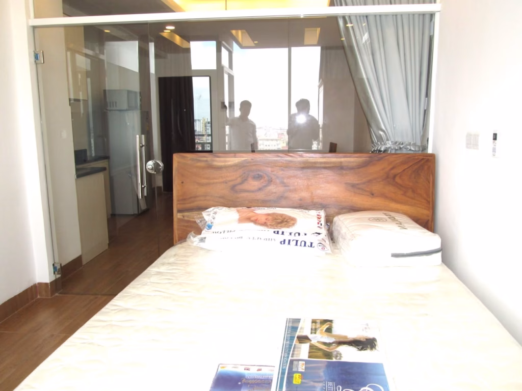 A modern bedroom in a studio apartment for sale features a wooden headboard, white bedding, and pillows. Reflected in the glass wall are two people and city views, while magazines rest on the bed.