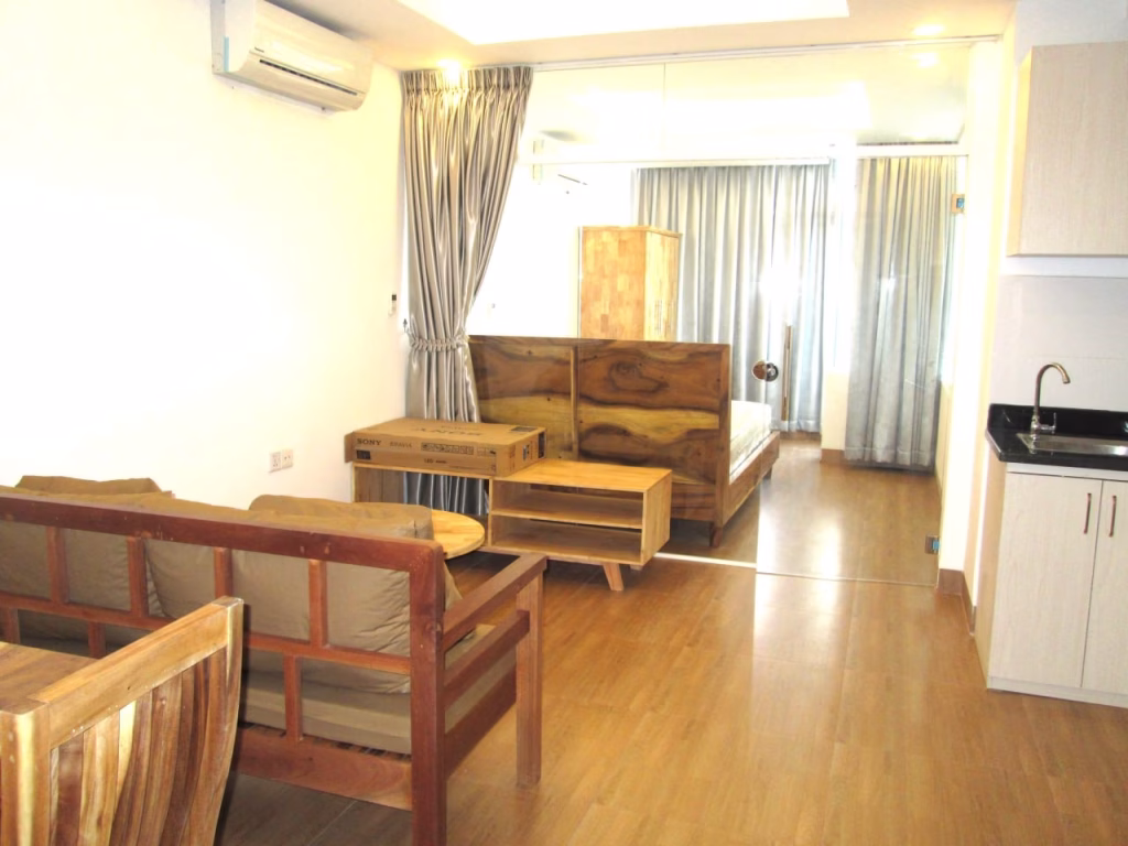 A modern studio apartment for sale with wooden furniture, including a sofa, tables, and bed. The space features wood flooring, neutral curtains, white walls, a small kitchenette, and an air conditioner on the wall.