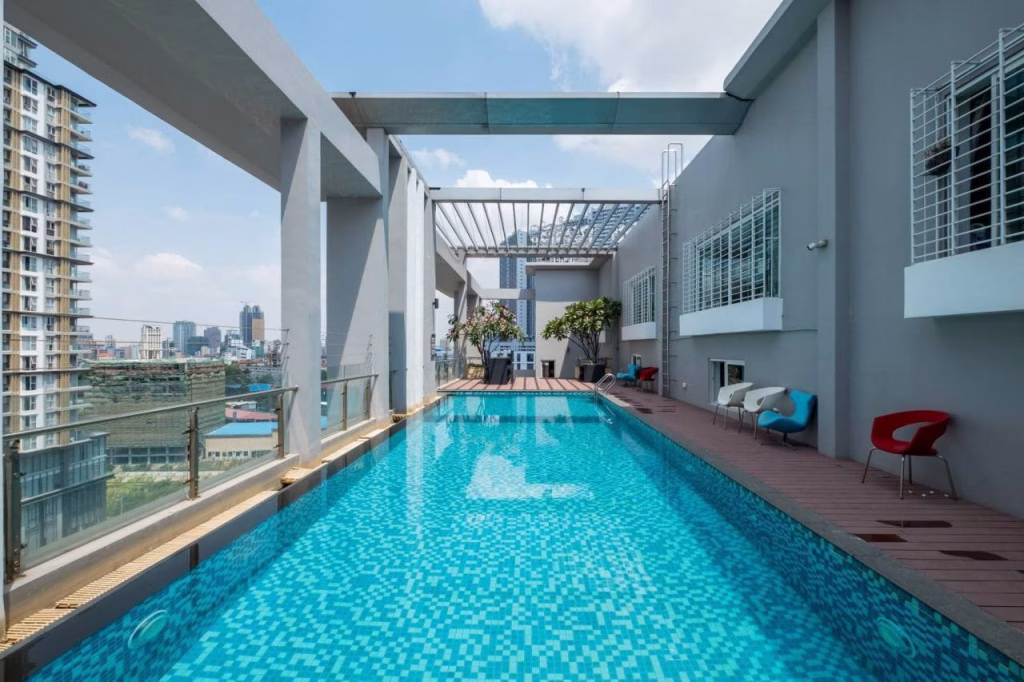 A modern rooftop swimming pool with blue tiles sits atop Diamond Island, surrounded by a wooden deck, lounge chairs, and potted plants. Tall buildings and a cityscape stretch out in the background under a partly cloudy sky.