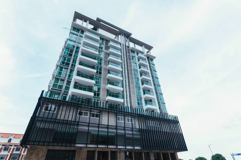 A modern multi-story building with glass balconies and large windows rises on Diamond Island, viewed from a low angle against a pale blue sky. The lower facade features striking vertical black slats.