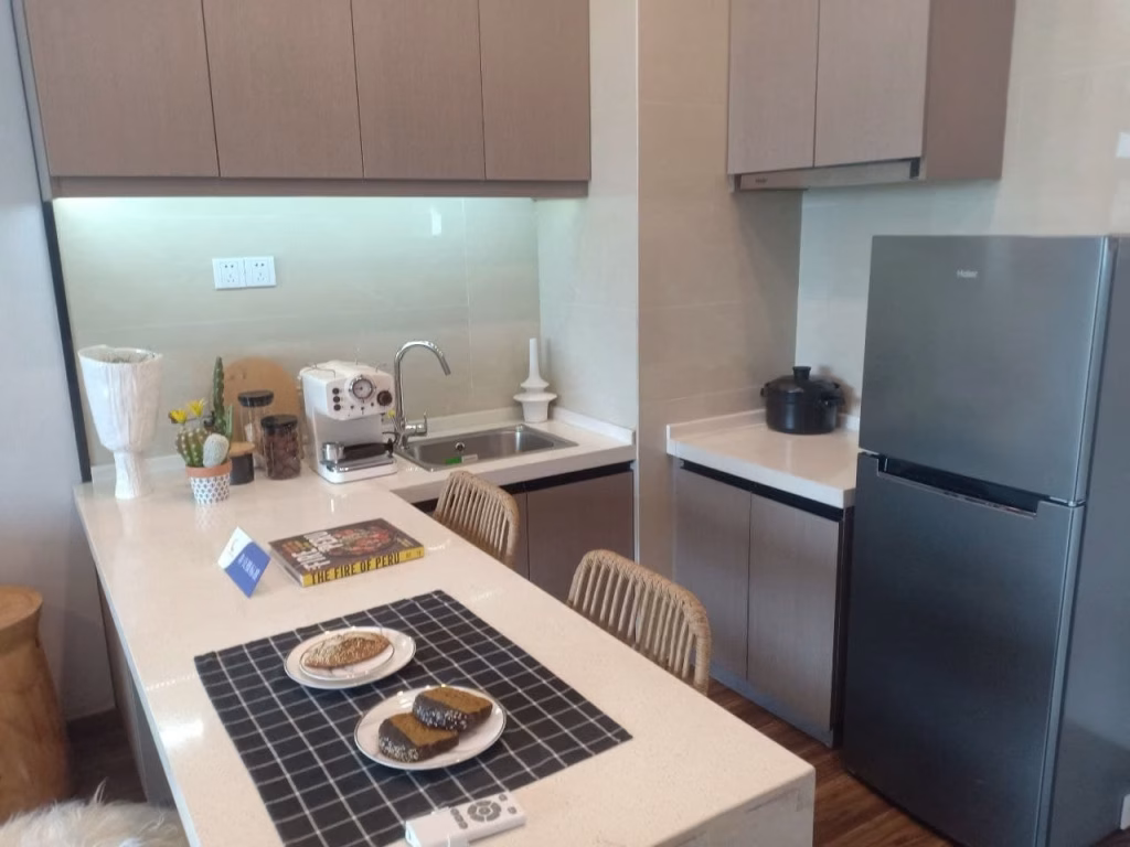 A modern kitchen in this Sihanoukville Studio For Sale features light brown cabinets, a gray fridge, and countertop appliances. The island boasts a black placemat, desserts, a book, two wicker chairs, plus elegant decor and a plant.