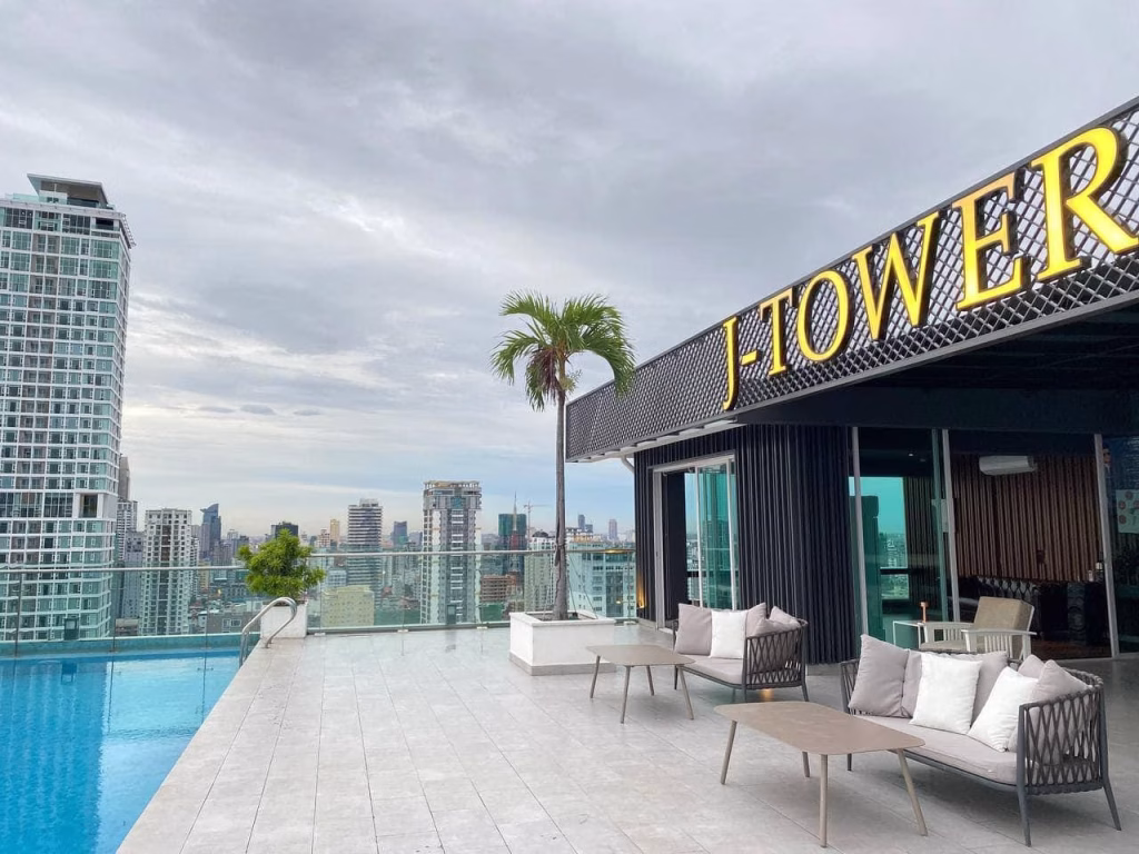 Rooftop pool area with lounge chairs and palm trees, overlooking a city skyline with tall buildings. A sign reading J-TOWER is visible on the right side of this Stunning Apartment For Sale. The sky is cloudy.