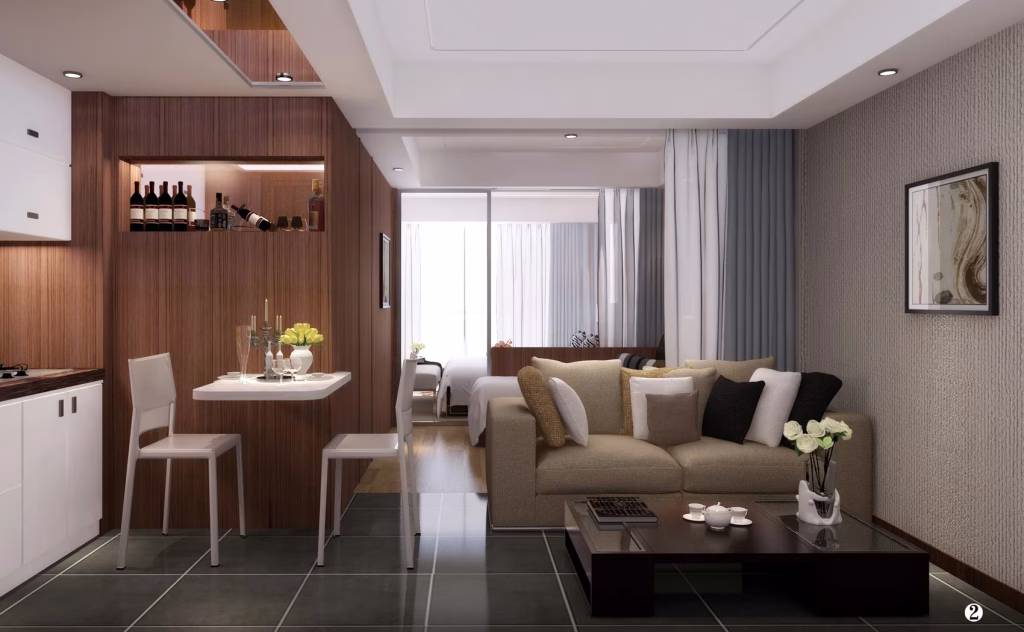 Modern studio at The Seagate Apartment with a kitchen, small dining area, and living space featuring a beige sofa, dark coffee table, and wall art. A partition separates the living area from a bedroom with large window and sheer curtains.