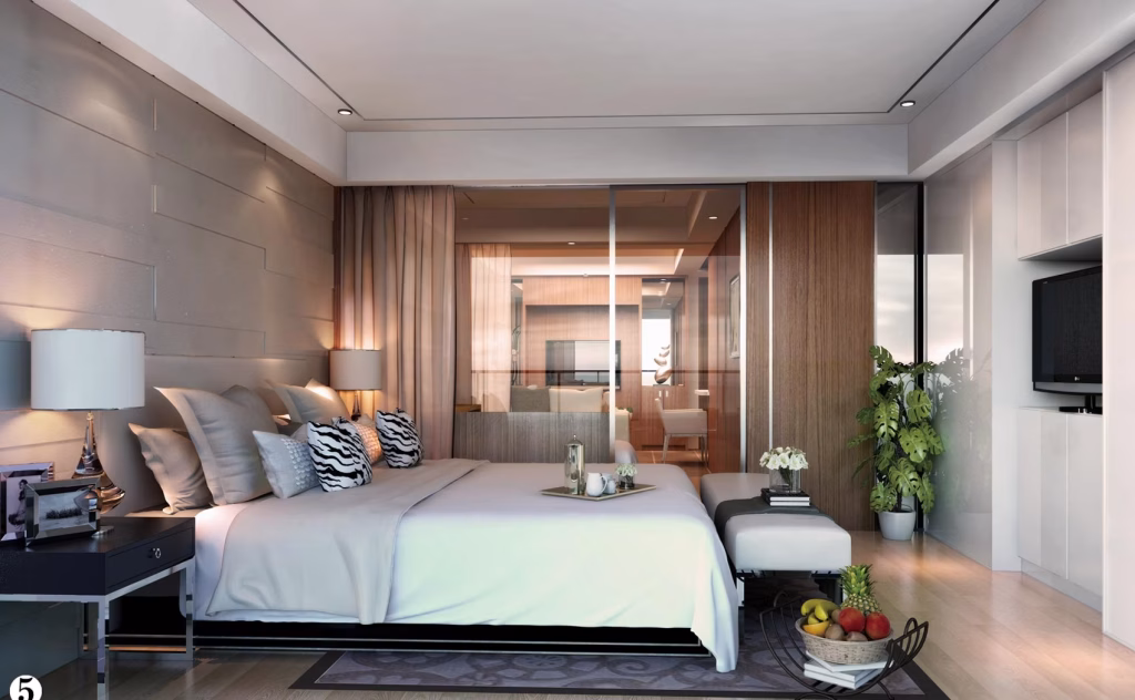 A modern bedroom at The Seagate Apartment features a large bed with decorative pillows, a nightstand with a lamp, a bench at the foot of the bed, indoor plants, a TV, and glass doors leading to a bright living area.