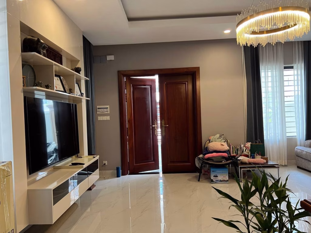 A modern living room in a twin villa for sale, featuring a TV unit on the left, large wooden double doors slightly open in the center, a round bench with folded clothes to the right, and a chandelier. Sunlight streams through the window.