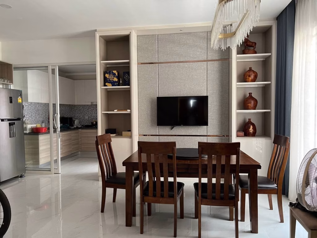 A modern dining area in this twin villa for sale features a wooden table and four chairs, a TV mounted on the wall, shelves with decorative vases, and a view into a kitchen with stainless steel appliances. Natural light fills the space through large windows.