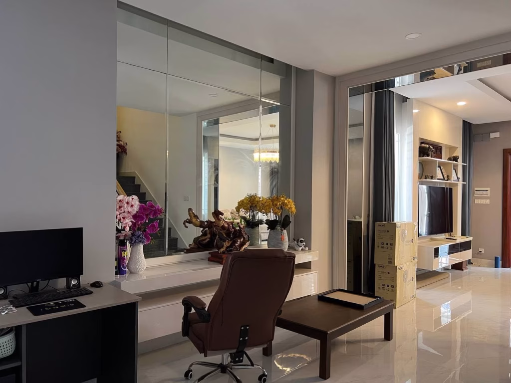 A modern living room in a twin villa for sale features a desk, office chair, computer, large mirrors, decorative flowers, and a TV on a wall unit. Cardboard boxes rest on glossy tiled floors with stairs visible in the background.