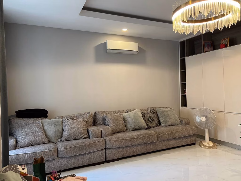 A modern living room in a twin villa for sale features a large gray sectional sofa, multiple cushions, wall-mounted air conditioner, standing fan, built-in shelves, and a decorative chandelier atop white glossy floors.