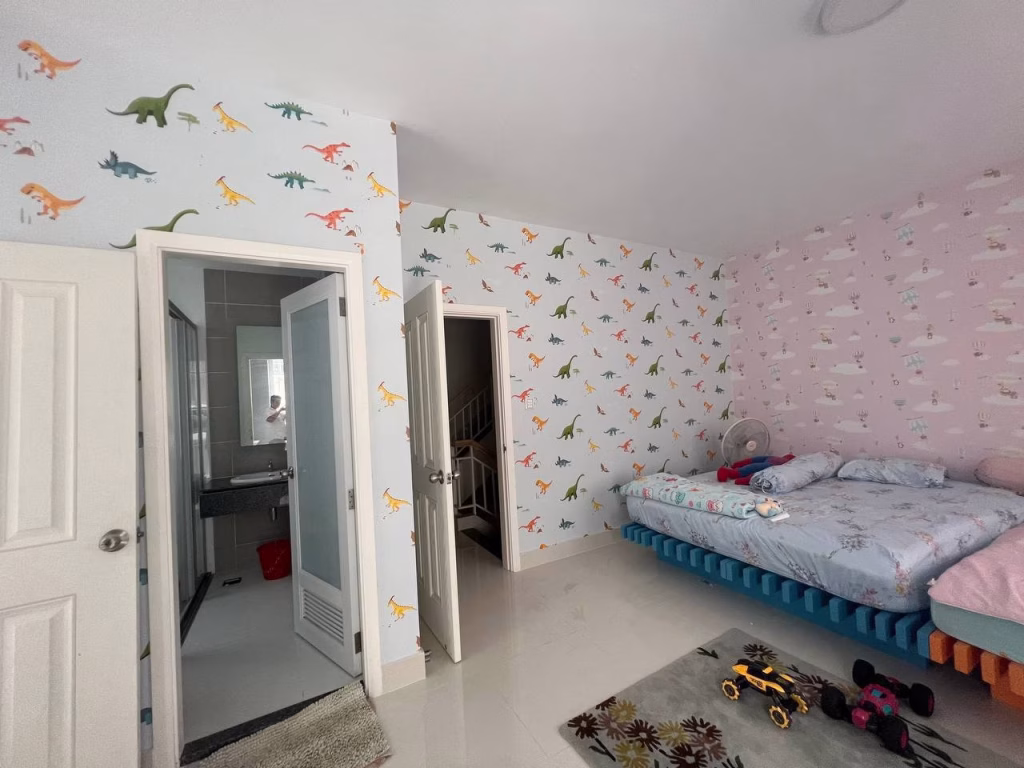 A childrens bedroom in a twin villa for sale features dinosaur and animal wallpaper, a blue bed with stuffed animals and a fan, a toy car, small rug, and open bathroom door. A staircase is visible through another door.