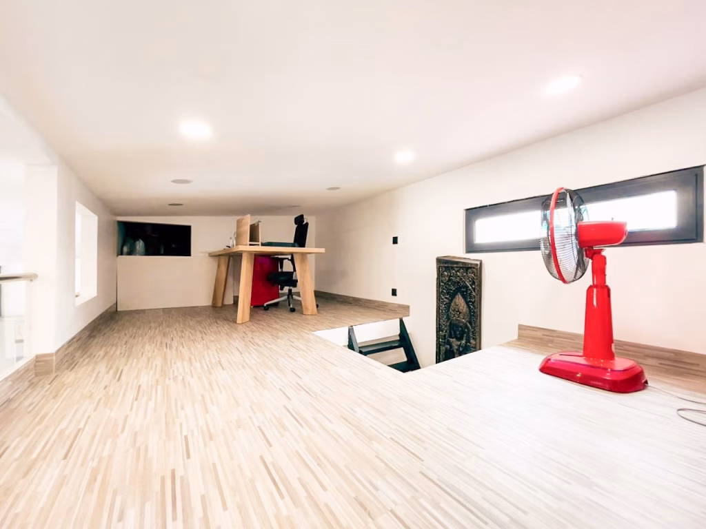 A minimalist attic workspace in a 3-bedroom apartment with a wooden desk, office chair, and large red floor fan, featuring light wood flooring and natural light from a rectangular window.