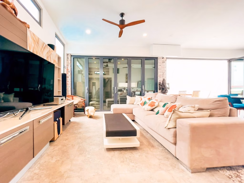 A modern living room in a 3-bedroom apartment features a large beige sectional sofa, dark rectangular coffee table, wall-mounted TV, and glass doors to a patio. Decorative pillows adorn the sofa, with a ceiling fan above. The space is bright and airy.