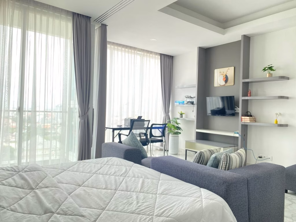 A bright, modern studio condominium at BKK1 with large windows, light curtains, a bed, gray sofa, wall-mounted TV, shelves with books and plants, and a small dining table with chairs near the window.