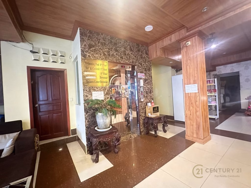 Hotel lobby with marble walls, decorative wooden furniture, potted plants, and an elevator. A dark wooden door sits to the left, vending fridge and shelves to the right. The Century 21 logo on the floor signals this hotel for sale.