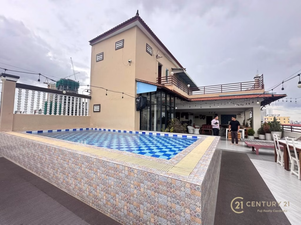 A rooftop terrace with a blue-tiled rectangular pool, outdoor dining area, string lights, and a beige two-story building. Two people stand near the entrance. The Century 21 logo indicates this hotel for sale opportunity.