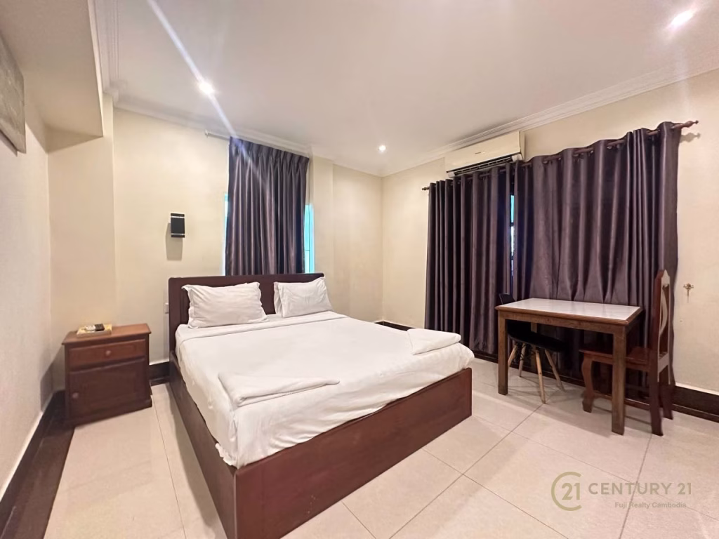 A neatly made double bed in a simple bedroom with beige walls, white tile floor, two windows with dark curtains, a wooden desk and chair, and a nightstand with a phone—perfect for showcasing this hotel for sale. Century 21 logo is visible in the corner.