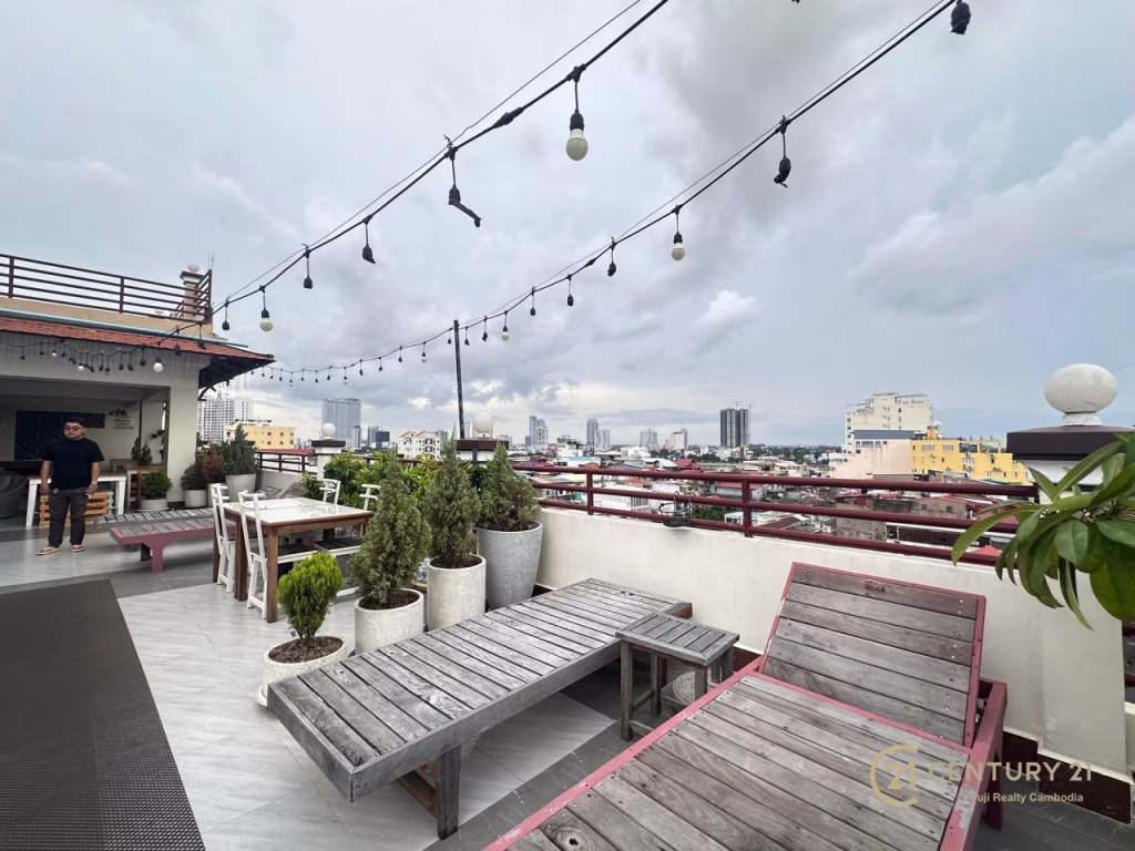 A rooftop terrace with wooden benches, potted plants, and string lights overlooks a city skyline under a cloudy sky. A person stands near the entrance on the left. Ideal for a boutique hotel for sale. The Century 21 logo is visible in the bottom right corner.