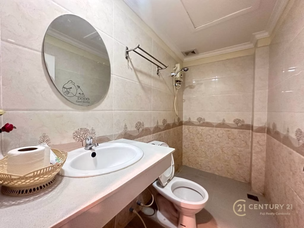 A small bathroom in a hotel for sale features a round mirror above a white sink, a toilet, wall-mounted shower, beige tiled walls with tree patterns, and a towel rack. A toilet paper roll and flower sit on the sink counter.