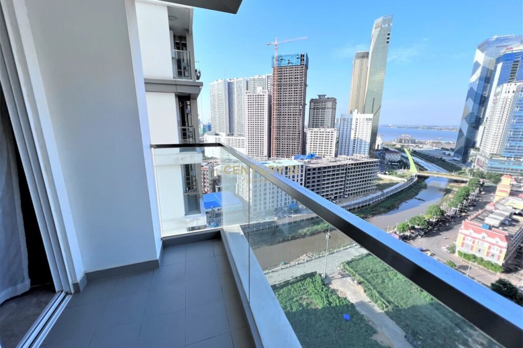 View from an Apartment 1 Bedroom high-rise balcony overlooking a cityscape with tall buildings, a river, construction sites, and green spaces under a clear blue sky.
