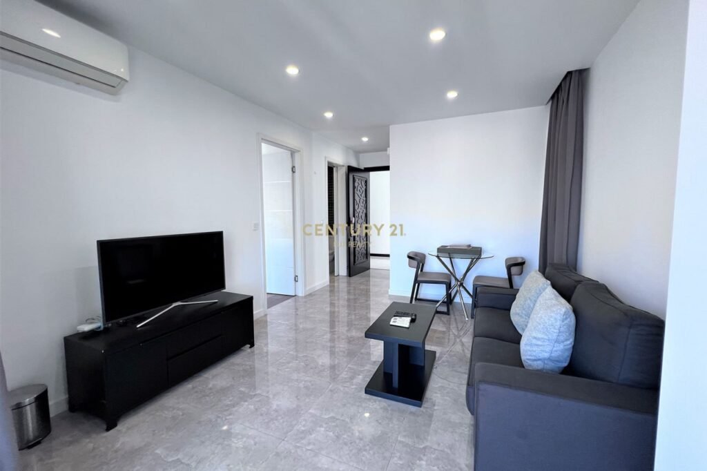 Modern 1 bedroom apartment living room with gray sofa, black coffee table, TV on a black stand, and small dining table for two. The space features tiled floors, white walls, recessed ceiling lights, and a door leading to another room.