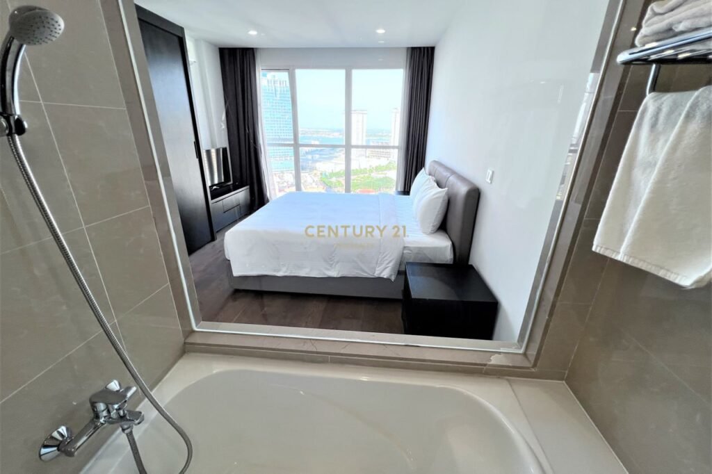 View from a bathroom with a bathtub looking into a modern Apartment 1 Bedroom featuring white bedding, bedside tables, and a large window showcasing cityscape and ocean views. Towel rack and CENTURY 21 logo in the center.